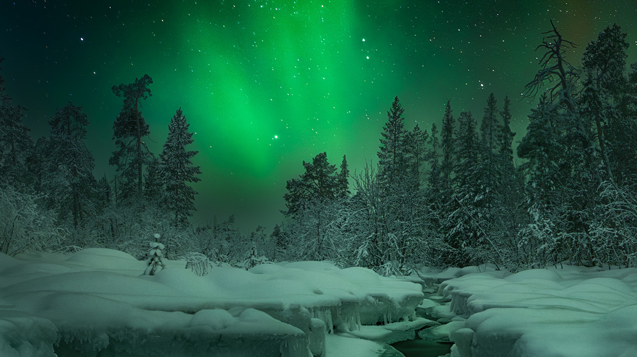Почти полностью скованное льдом русло северной реки отражает зеленые всполохи полярного сияния. Мурманская область.